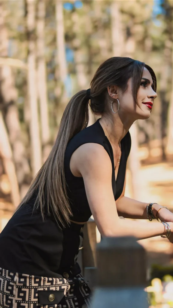 A side profile of a woman with long, ash-blonde and brown hair styled in a sleek, low ponytail secured at the nape of the neck. Her hair is straight and extends down her back, with a single face-framing strand left loose. She is wearing a black sleeveless top, silver hoop earrings, and bold red lipstick, leaning against a wooden railing. The background features a sunlit, blurred forest with tall, slender trees.
