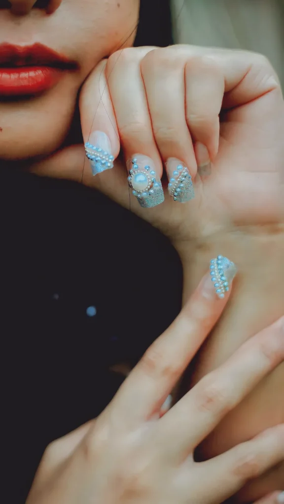 A close-up of a glamorous manicure featuring long, square nails adorned with intricate 3D embellishments. The design includes a mix of shimmering silver glitter, small blue rhinestones, and delicate white pearls arranged in textured patterns. One accent nail features a large, central statement pearl surrounded by a halo of tiny gems. The hands are posed elegantly near the face, highlighting the "fiercely red-carpet-ready" drama of the 3D art.