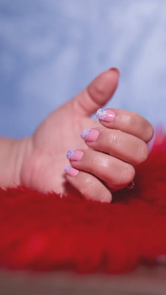 A close-up of a hand resting on a vibrant red faux-fur surface. The short, square-shaped nails feature a soft bubblegum pink base with a shimmering, iridescent "glazed" finish on the tips. The tips sparkle with fine purple and blue glitter, catching the light to create a glowing, ethereal effect. The background is a soft-focus blue, emphasizing the "mesmerizing, light-catching finish" of the manicure.