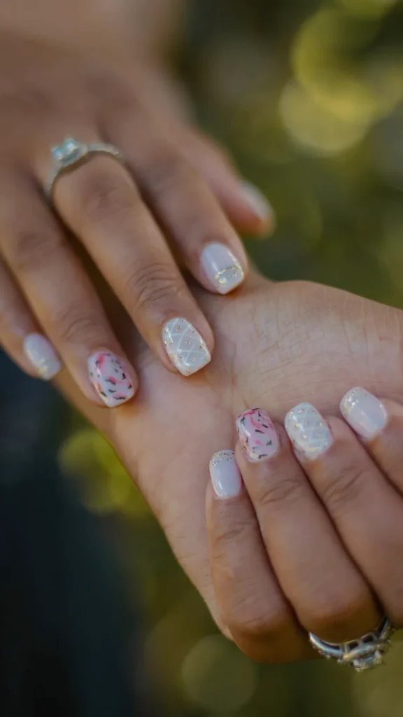 A close-up of hands featuring a highly personalized, abstract manicure on short, square-shaped nails. The set includes a mix of artistic designs: soft white bases with gold glitter stripes, a delicate white lattice pattern with gold accents, and a standout abstract nail with swirling brushstrokes of pink, black, and gold glitter over a sheer base. The hands are held together against a soft, natural green background, showcasing the unique and "highly personalized" vibe.