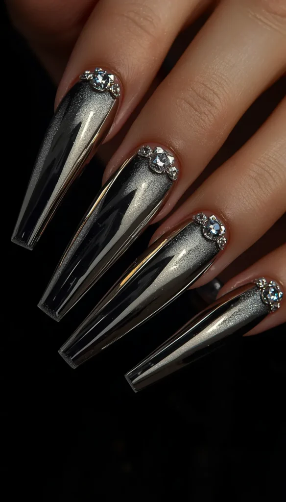 A close-up view of a hand displaying long, coffin-shaped nails against a stark black background. The nails feature a highly reflective, dark silver and black metallic chrome finish, with a bright, luminous streak down the center resembling a cat-eye polish effect. At the cuticle base of each nail, there is a decorative cluster of clear, sparkling rhinestones consisting of a larger central gem flanked by smaller crystals.