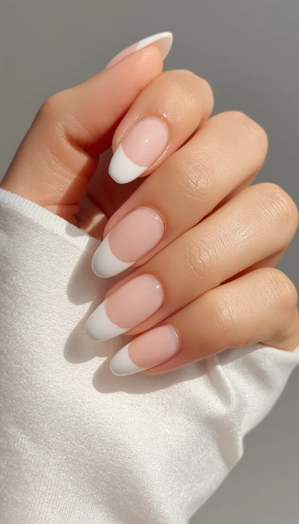 A close-up of a hand resting gently against a smooth, white fabric sleeve, illuminated by soft natural sunlight. The hand displays a fresh, glossy manicure with almond-shaped nails. The design is a classic, minimalist French tip, featuring a sheer, natural pink base with clean, curved white tips. A plain, light gray background is visible behind the hand.