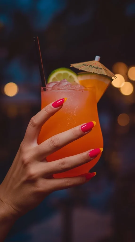 ALT Text:
A close-up shot of a hand holding a tall, frosted glass of a vibrant orange tropical cocktail filled with crushed ice. The cocktail is garnished with a lime wheel, an orange slice, a black straw, and a small paper cocktail umbrella. The person's long, almond-shaped nails feature a bold hot pink manicure tipped with a bright yellow-orange accent, reminiscent of a sunset. The background is a moody, dim evening setting with warm, golden bokeh lights creating a festive nightlife ambiance.